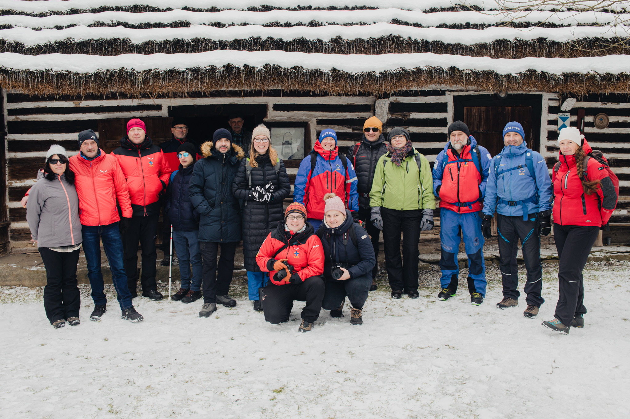 Photo of 16 people wearing winter sport clothes, standing in front of the wooden shelter hut covered by snow. Cheerful atmosphere, training participants are smiling.