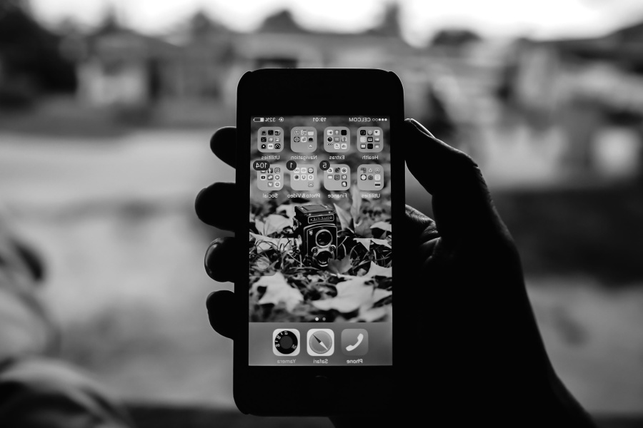 Czarno-białe zdjęcie. Prawa dłoń trzymająca smartfon. Na jego ekranie widać wiele folderów zawierających różne aplikacje mobilne.